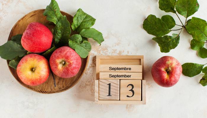 Four red apples and green leaves surround a wooden calendar displaying September 13, at the height of apple-picking season in Quebec.